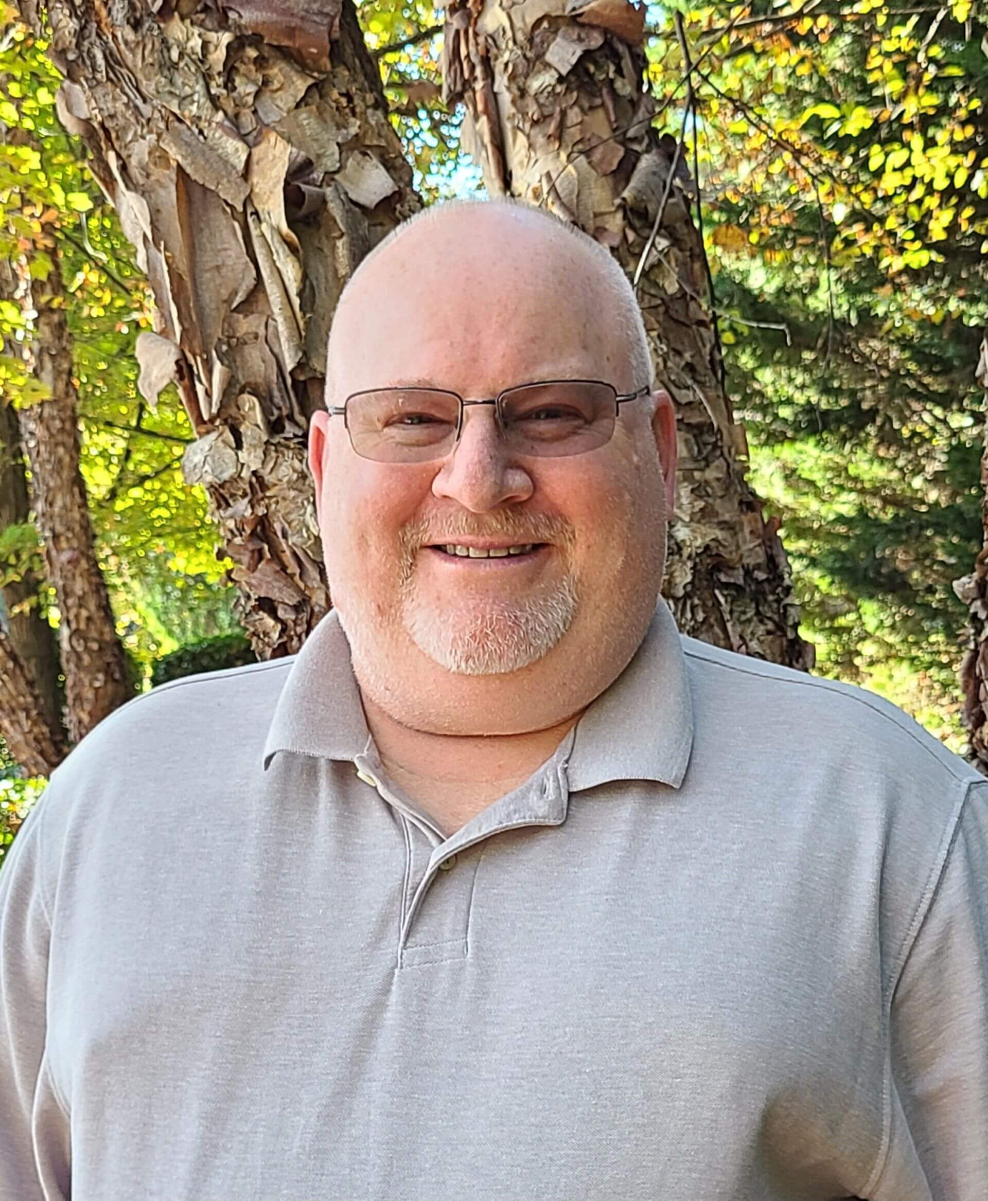A bald man smiling in front of a tree.